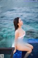 A woman in a white bathing suit sitting on a blue bench by the water.